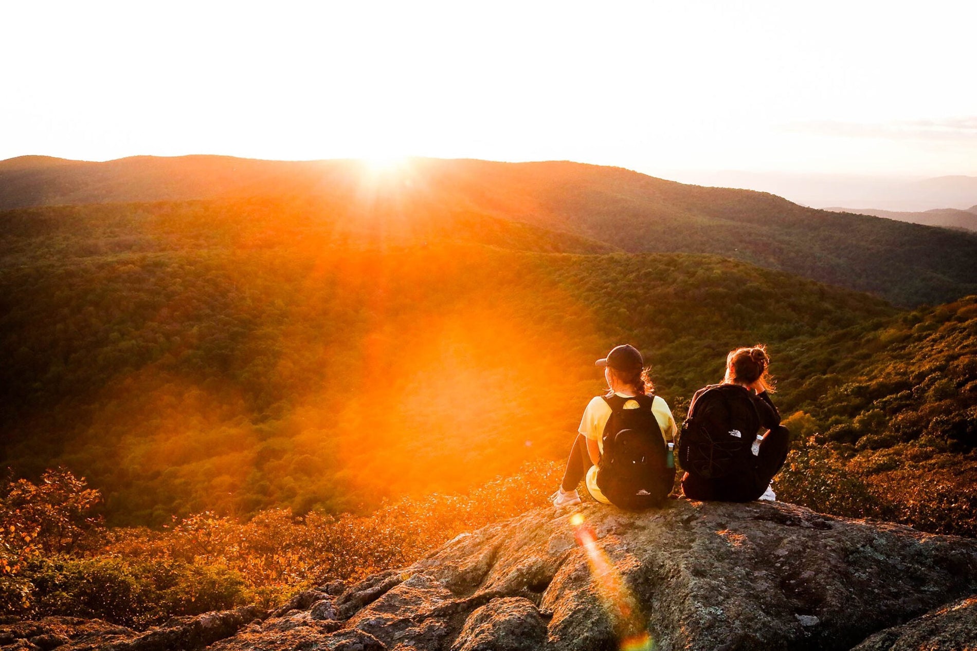 Two people sitting on a rock overlooking a scenic view of mountains during sunset.