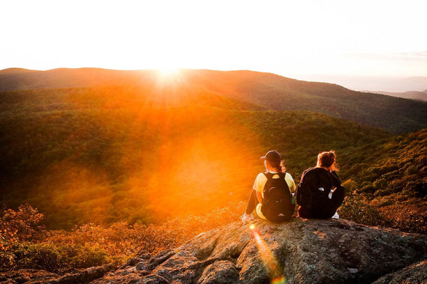 Two people sitting on a rock overlooking a scenic view of mountains during sunset.