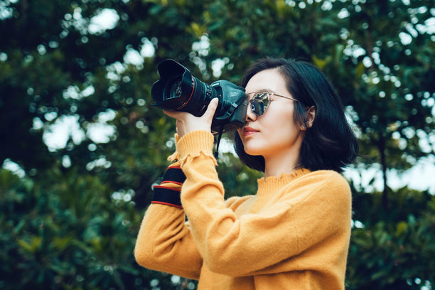 A person wearing a yellow sweater is taking a photograph with a camera, standing against a backdrop of green trees.